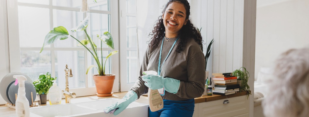 Selective focus on happy african american volunteer helping senior lady to clean house, standing in kitchen in raisin gloves washing sink with detergent, talking to elderly female sitting at table